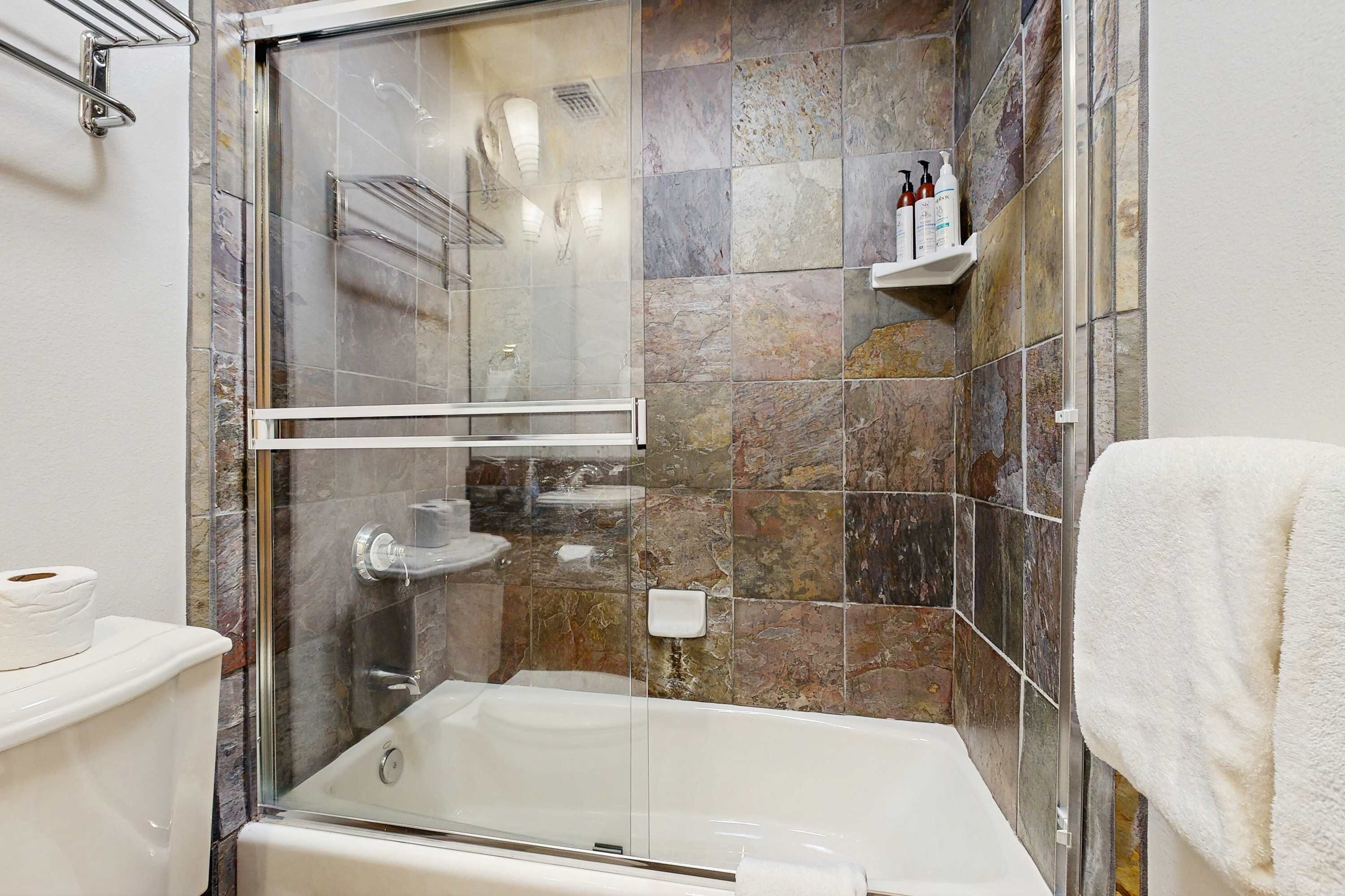 A bathroom with a glass-enclosed shower, a bathtub, a toilet, and a shelf with toiletries.