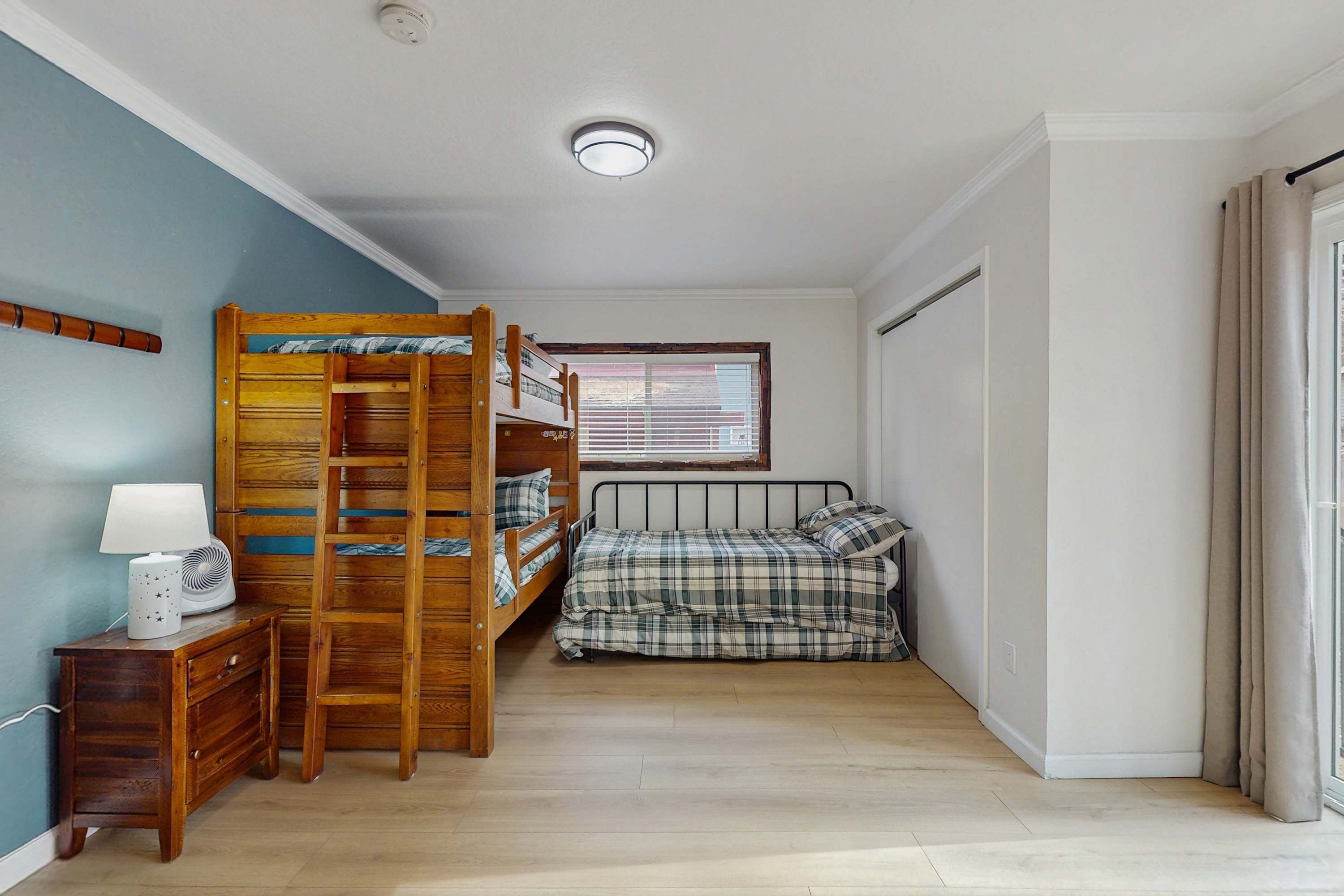 Une chambre avec un lit superposé, une table de chevet avec une lampe, une fenêtre avec des stores et une porte.