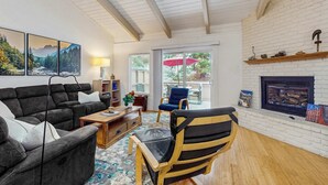 Een woonkamer met een bank, fauteuil, bijzettafeltje, boekenkast, open haard en uitzicht door een glazen deur.