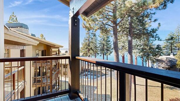 A wooden deck with a metal railing overlooking a building with a staircase and a view of trees and a body of water. - Comfortable Townhome with Pool, Deck, & Lovely Sunsets - Walk to Lake (Kings Beach)