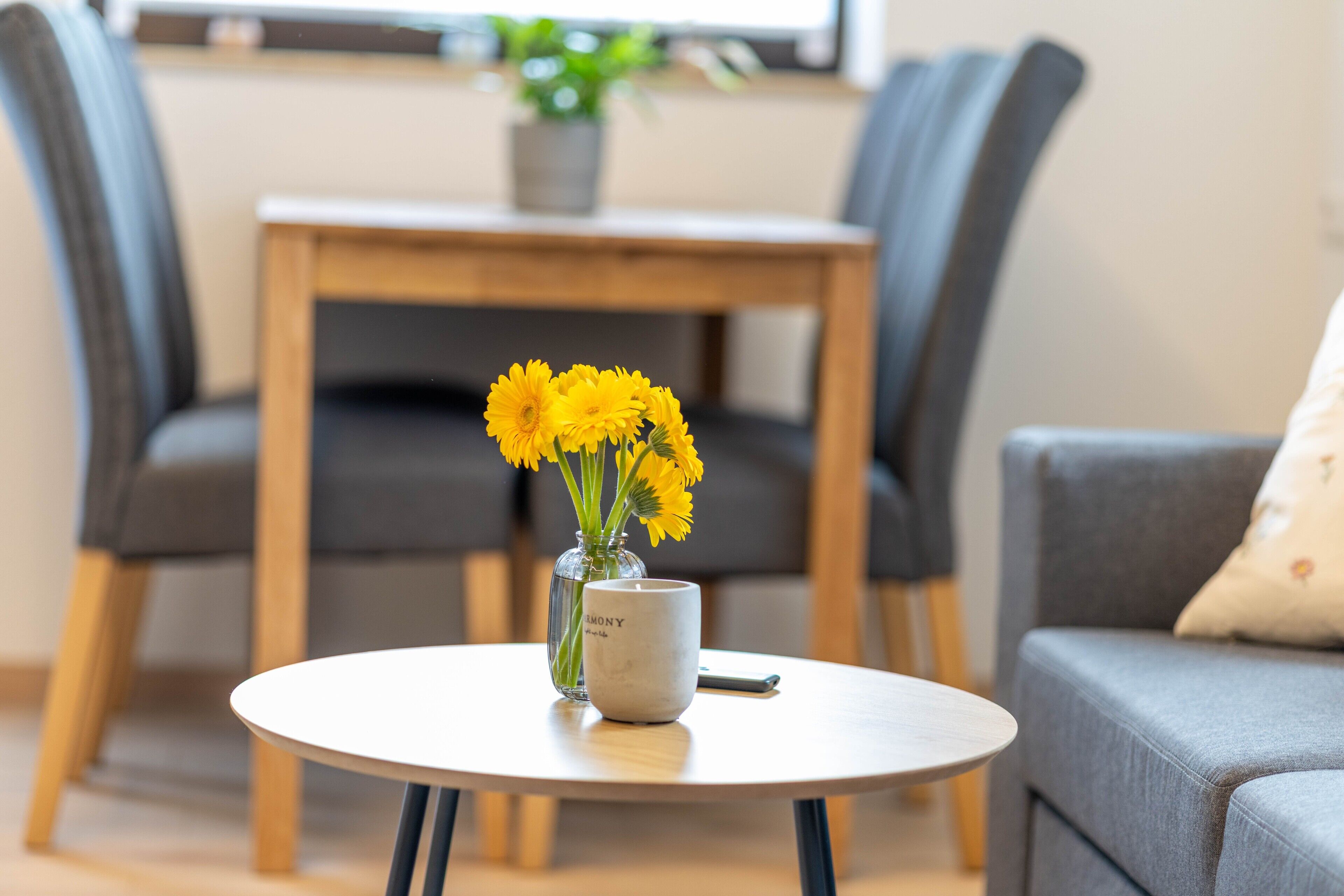 Ein runder Tisch mit einer Vase voller gelber Blumen und einer Tasse, ein graues Sofa und ein Holzstuhl in einem Zimmer mit einem Fenster und einer Pflanze.