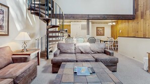 Un vaste espace de vie comprenant un canapé en cuir marron, une table basse avec un magazine, un escalier en colimaçon et, au fond, un coin repas.