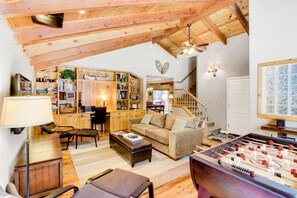 Een gezellige woonkamer met een bank, salontafel en een tafelvoetbalspel. Er is een boekenkast vol boeken en decoratieve objecten, en een trap die naar een ander niveau leidt.