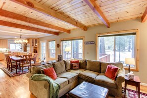 Ein gemütliches Wohnzimmer mit einer Eckcouch, einem Essbereich mit Holztisch und -stühlen sowie großen Fenstern mit Blick ins Freie.