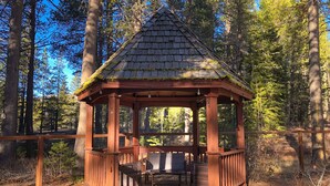 A wooden gazebo with a tiled roof, surrounded by a wooden deck and benches, set in a forested area. - Beautiful home with private hot tub, gazebo, and gas fireplace (South Lake Tahoe)