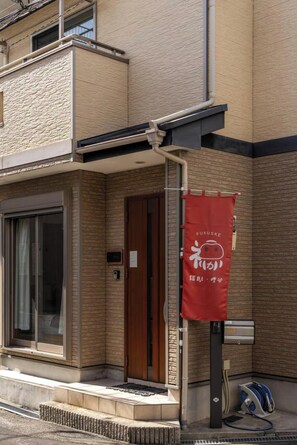Un bâtiment à la façade en briques, une banderole rouge avec du texte en japonais, une porte coulissante en verre et une petite marche menant à l’entrée.
