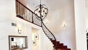 Eine Treppe mit hölzerner Handlauf und einer Chandelier, eine weiße Wand mit gerahmten Bildern, ein beiges Sofa mit gemusterten Kissen und ein Holzboden.