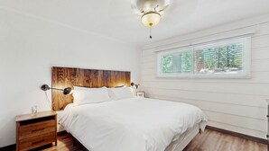 Ein Schlafzimmer mit Holz-Kopfteil, einem Nachttisch mit Lampe, einem Fenster mit Jalousien und einem Deckenventilator.