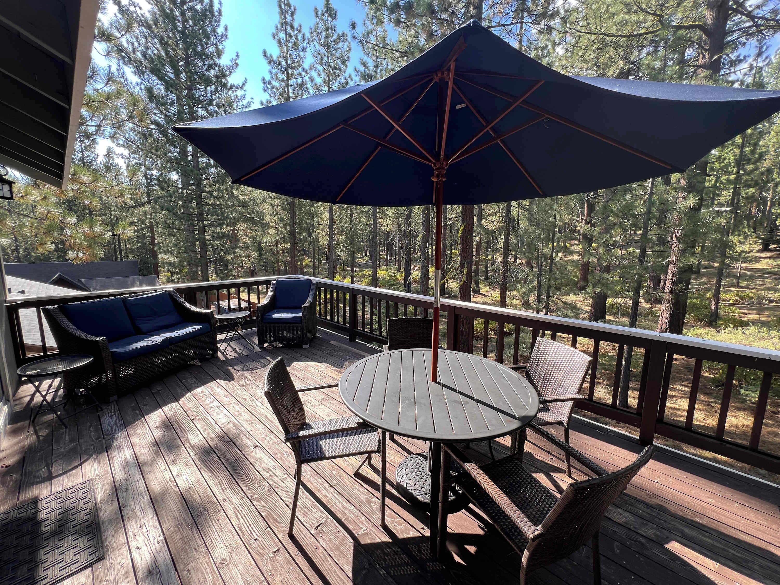 Um deck de madeira com móveis de exterior, incluindo uma mesa e cadeiras, e um grande guarda-sol a dar sombra.