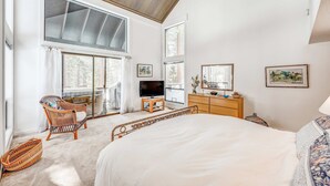 A bedroom with a large bed, a television, a wooden dresser, wicker chairs, and large windows. - Golf-course-front home with great deck, game room, fireplace & forest views (Incline Village)