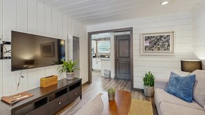 Een moderne woonkamer met een flatscreen televisie, een donker houten tv-meubel, een beige bank en een zichtbare keuken door een open deur.