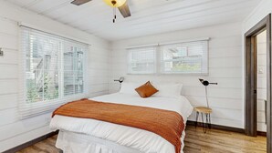 Een slaapkamer met een bed, een plafondventilator, twee ramen, een klein krukje en een deur.