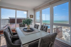 Uma sala de jantar com mesa de vidro, cadeiras acolchoadas, um vaso com folhagem e vista para o oceano e a cidade através de grandes janelas.