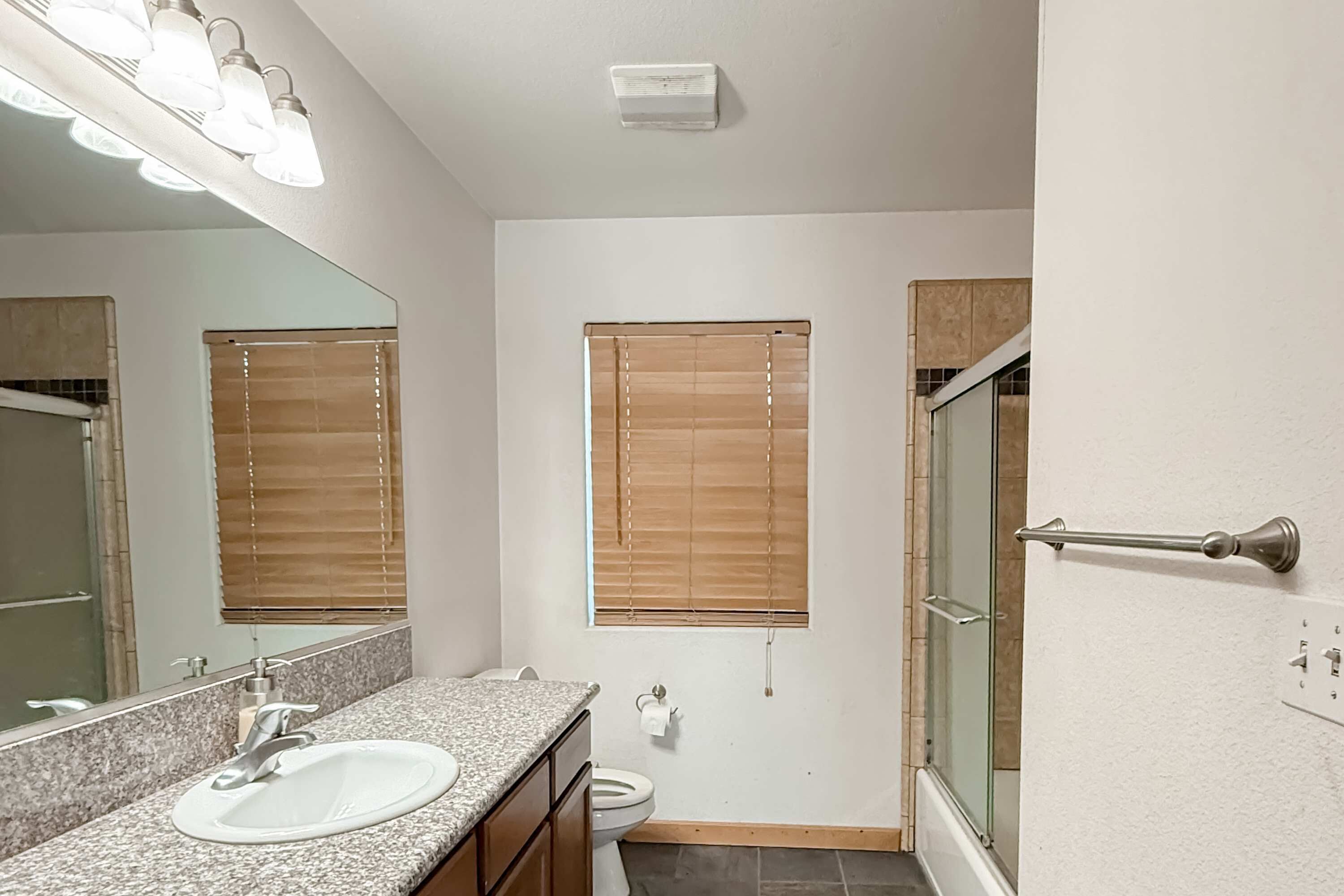 Un baño con encimera de granito, un lavabo blanco, un espejo, un inodoro y una ducha con puerta de cristal.