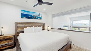 A bedroom with a large bed, a ceiling fan, a painting of a landscape, a nightstand, and a window with a view. - Luxury Condo in Stateline: Lake & Mountain Views (Stateline)