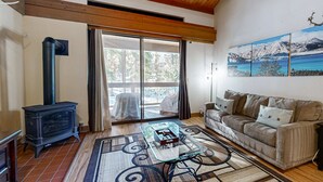 Una sala de estar con sofá, una mesa de centro de vidrio, chimenea y un ventanal con vista a árboles y montañas.
