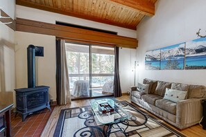 A living room with a sofa, a glass coffee table, a fireplace, and a large window with a view of trees and mountains. - Beautiful condo with shared pool, hot tub, & sauna - walking distance to skiing (Northstar - Truckee)