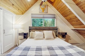 A bedroom with a sloped ceiling, a large bed with pillows, a nightstand, a lamp, and a window with a view of trees. - Chalet-style Townhome with Fireplace, Deck, Near Ski Resort, Incline Village (Incline Village)