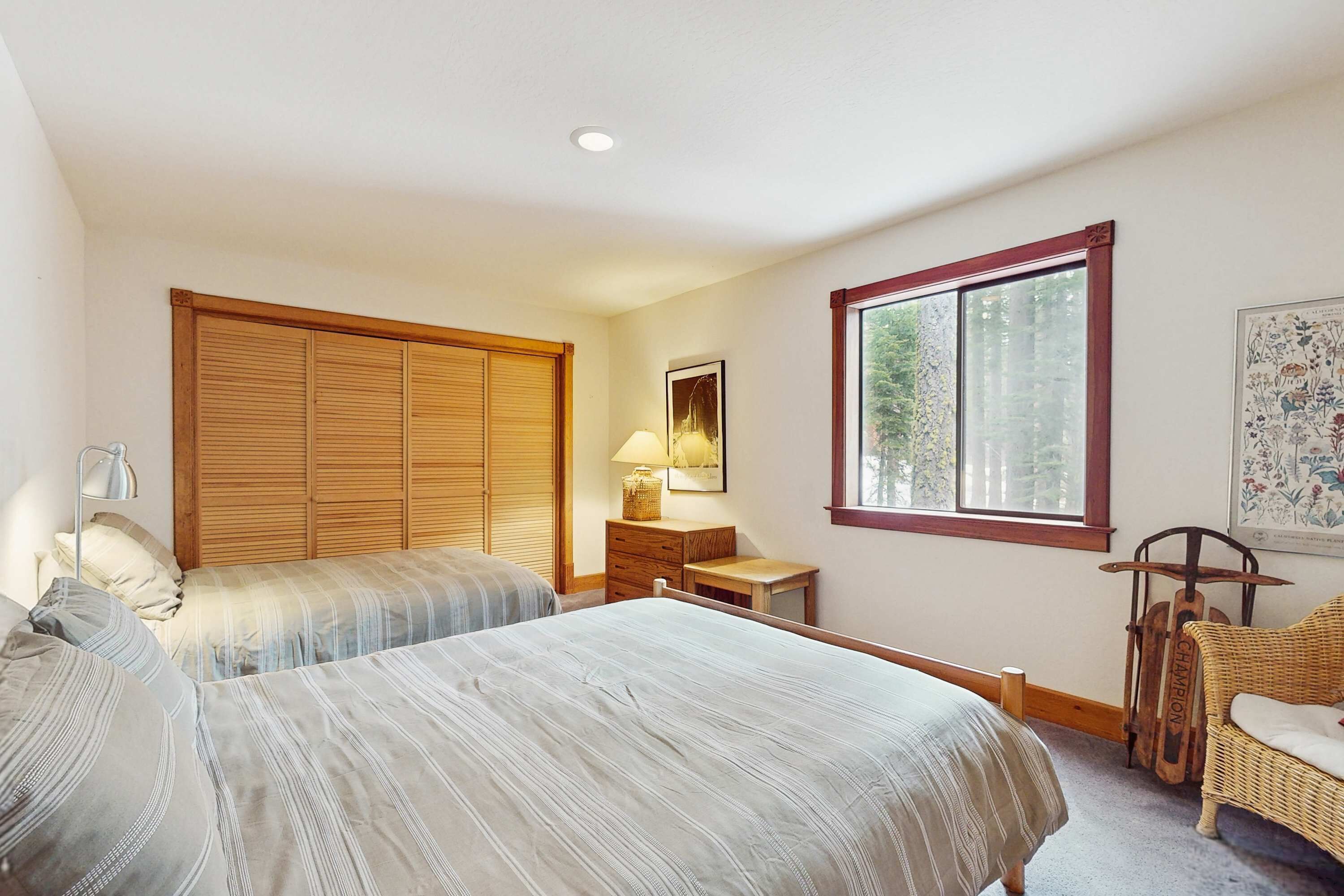 Un dormitorio con dos camas, un armario de madera, un escritorio con lámpara, una silla de mimbre y un ventanal grande.