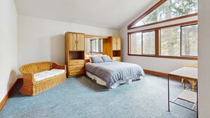 Un dormitorio con una cama, una silla de mimbre, un armario de madera y un ventanal grande.