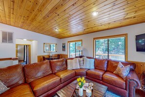 A living room with a wooden ceiling, a brown leather sectional sofa, a coffee table with a vase of flowers, and large windows. - Lovely mountain home with skiing, pool, hot tub, fast WiFi, & washer&dryer (Truckee)