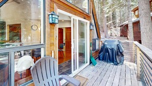 A wooden cabin with a deck, a covered outdoor seating area, and a large window overlooking the forest.