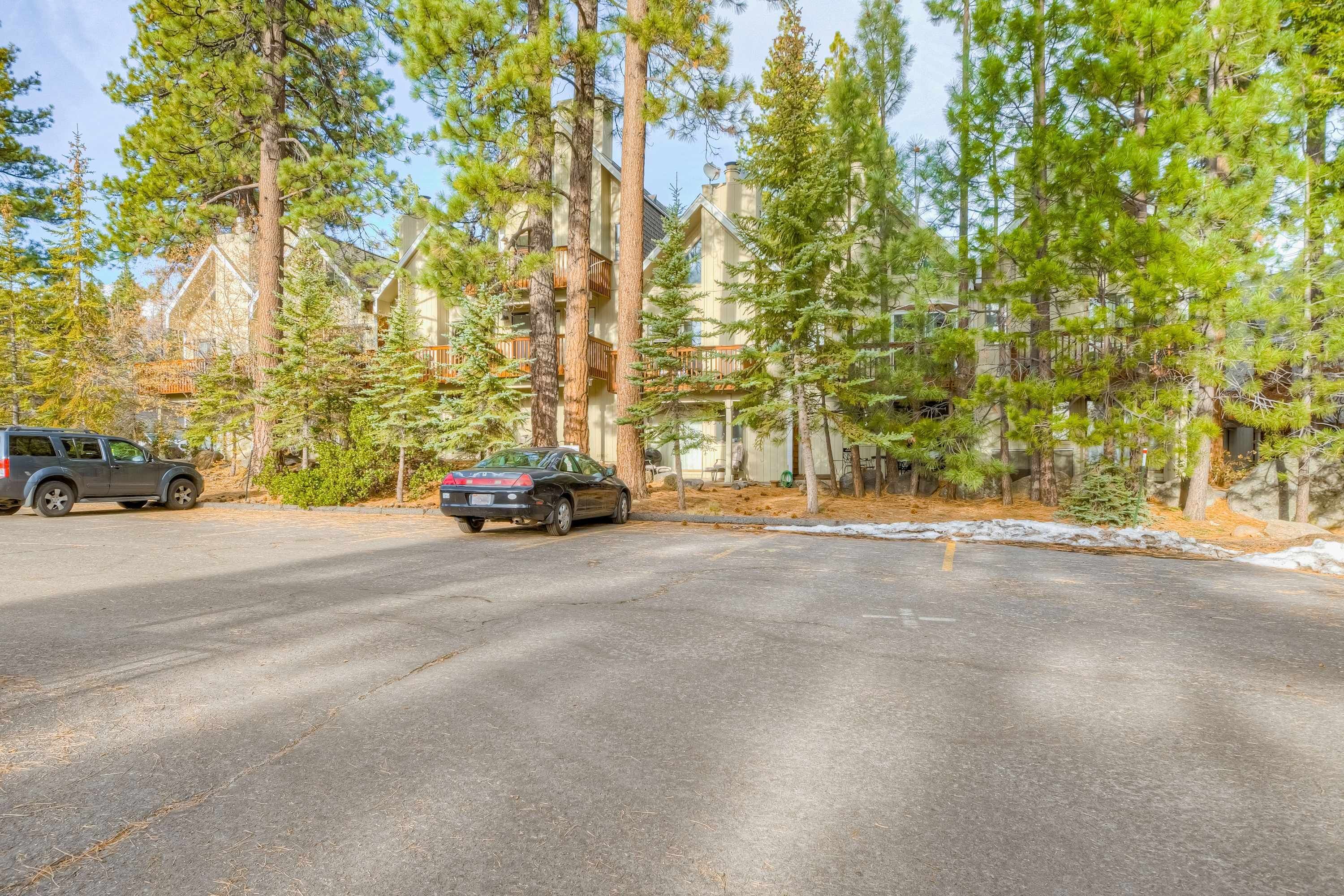 A road with two cars parked on the side, surrounded by tall pine trees and a building with a balcony.