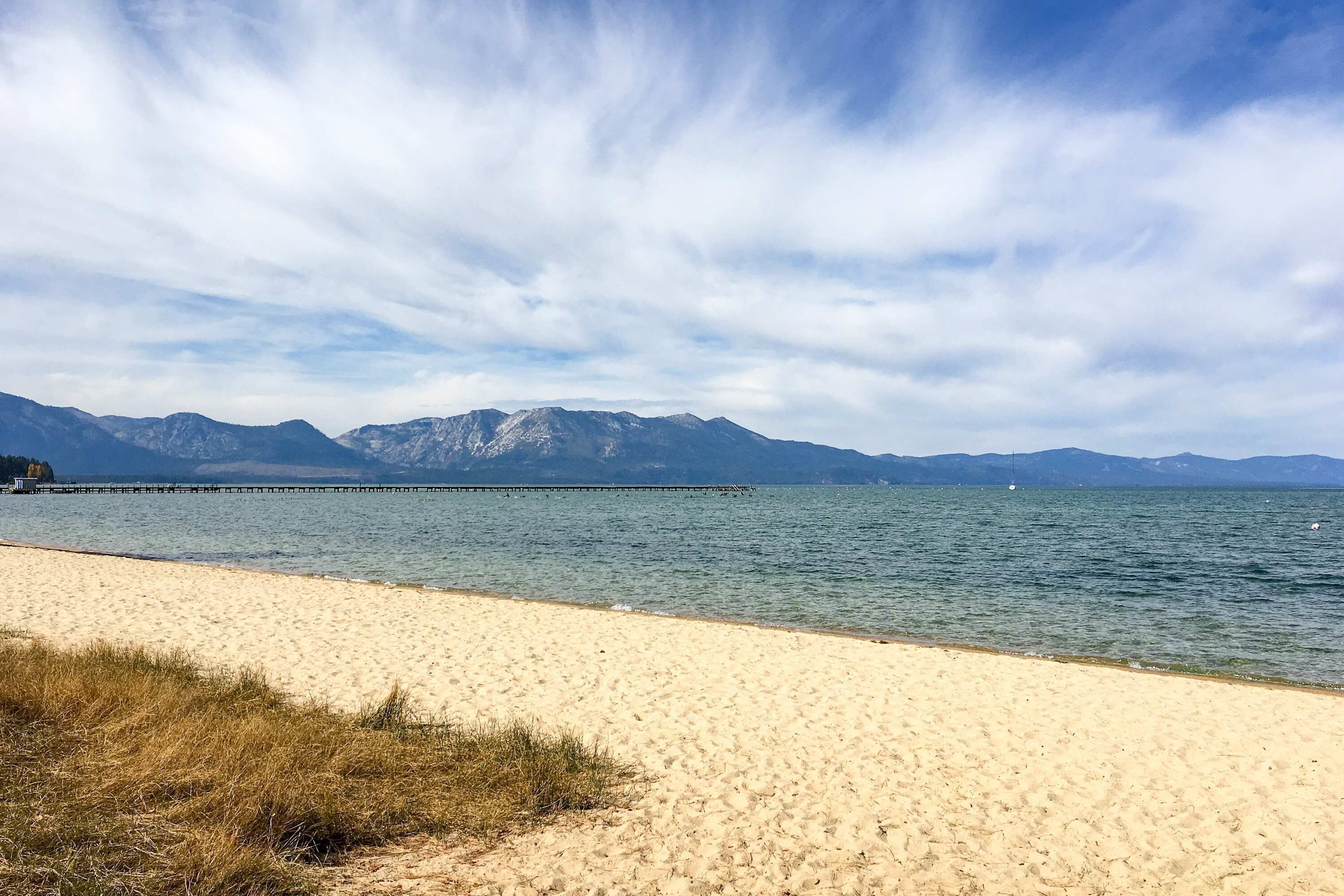 Une plage avec un ciel dégagé, des montagnes en arrière-plan et une jetée qui s’avance dans l’eau.