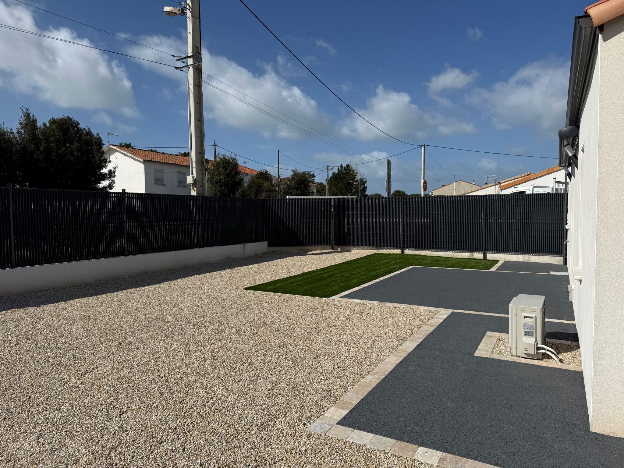 Un espace de stationnement avec une surface en gravier, une partie goudronnée et une clôture noire.