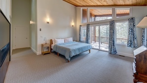 Une chambre à coucher comprenant un lit, une table de chevet, une lampe, une commode et de grandes fenêtres avec des rideaux.