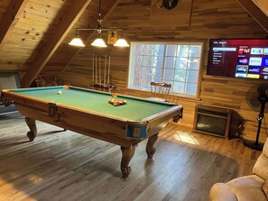 A pool table in a room with wooden walls, a large window, and a mounted TV displaying various channels. - Inviting home with pool table - near Heavenly & beaches (South Lake Tahoe)