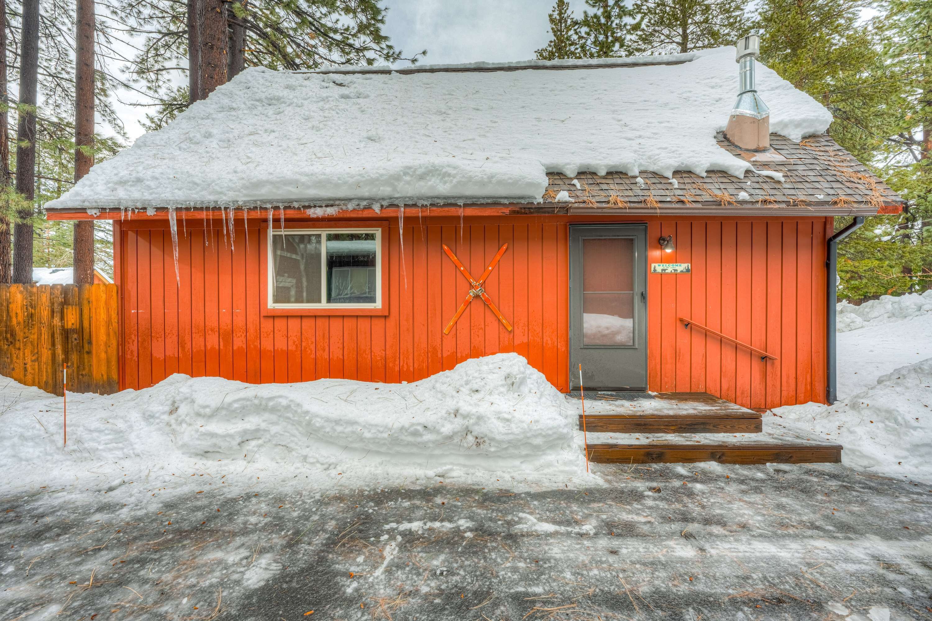 Une petite cabane en bois rouge, avec un toit enneigé, une cheminée et une porte munie d’une main courante.