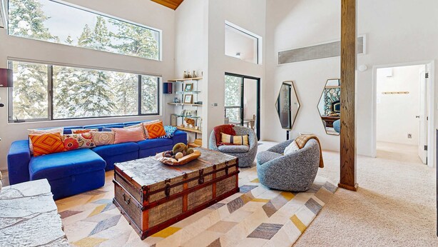 A spacious living area with a blue sofa, a grey armchair, a wooden chest, and a large window offering a view of trees. - Modern mountain home with private hot tub, deck, fireplace, & skiing nearby (Northstar - Truckee)