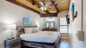 Une chambre avec un plafond en bois, un lit recouvert de linge de lit blanc, une table de chevet avec une lampe, un miroir et des œuvres d’art accrochées aux murs.