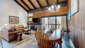 Un vaste salon doté d’un plafond en bois, d’une cheminée, d’un téléviseur et d’un coin repas avec des chaises et une table en bois.