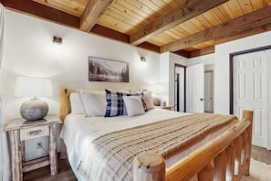 A bedroom with a wooden ceiling, a bed with a patterned blanket, a painting on the wall, and a nightstand with a lamp. - Delightful South Lake Tahoe Cabin with BBQ, Decks, Fireplace & Hot Tub - Minutes to Beach&Ski (South Lake Tahoe)