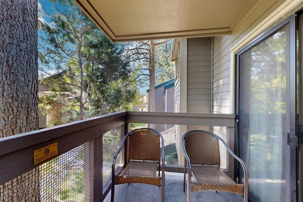 A covered balcony with two wicker chairs, a mesh safety barrier, and a view of trees and buildings. - Three-Story Condo & Loft w Free WiFi, Gas Fireplace, Shared Pool, Hot Tub, & WD (South Lake Tahoe)