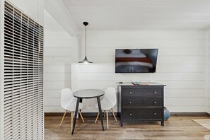 Ein Zimmer mit einem Esstisch, zwei Stühlen, einer schwarzen Kommode, einem an der Wand befestigten Fernseher und einer Deckenleuchte.