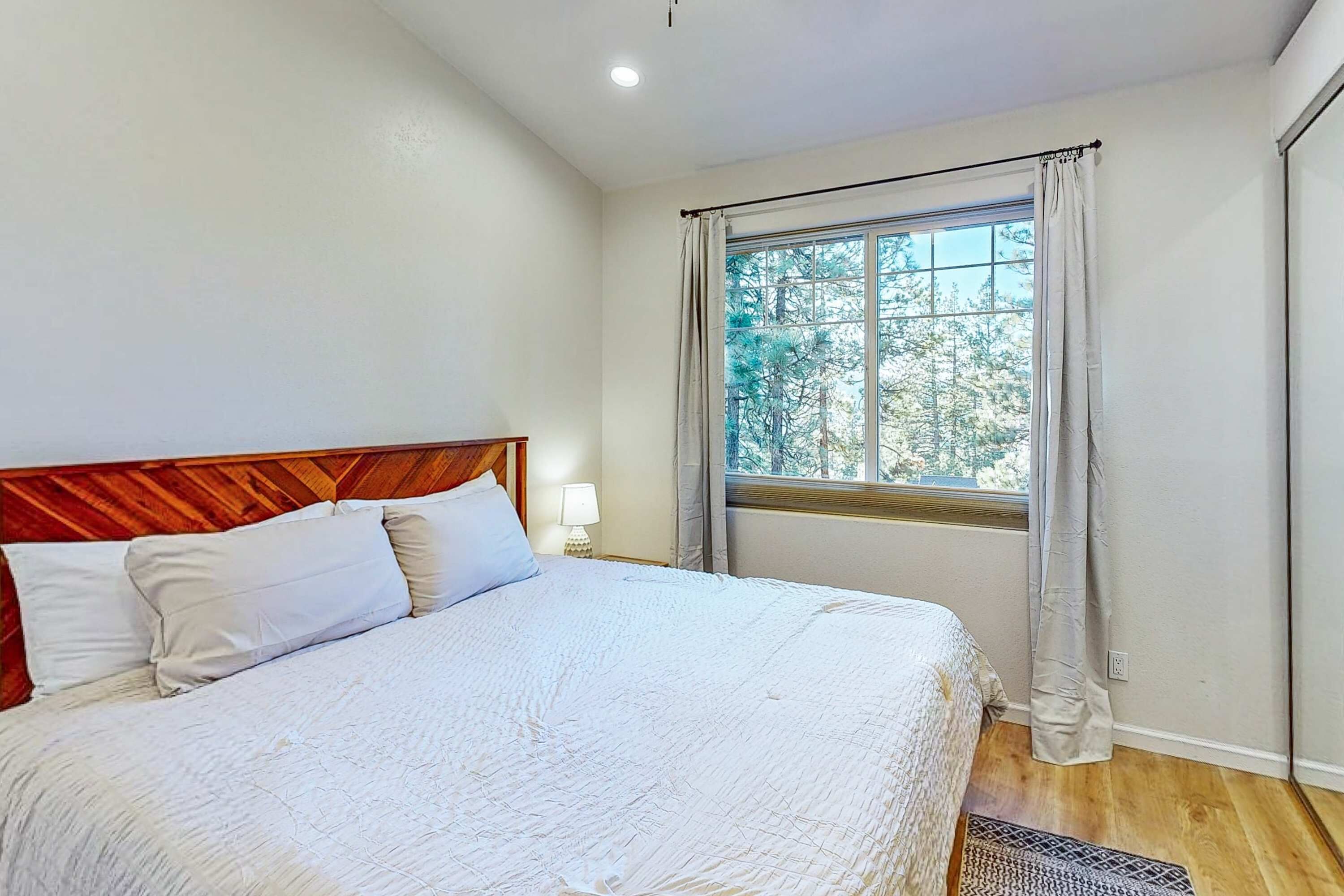 Un dormitorio con cabecera de madera, una cama grande con ropa blanca, una ventana con cortinas y una mesita de noche con lámpara.