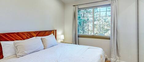 Un dormitorio con cabecera de madera, una cama grande con ropa blanca, una ventana con cortinas y una mesita de noche con lámpara.