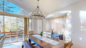 Un comedor con mesa de madera, sillas, una araña y ventanas grandes con vista a árboles.