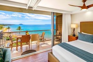 Een balkon met uitzicht op de oceaan, een bed met een blauw kussen en een houten nachtkastje met een lamp.