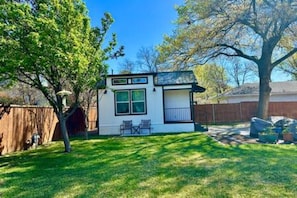 A small, single-story building with a white exterior and a black roof, surrounded by a wooden fence and trees. - New Cottage + Monthly + Fire Pit + Outdoor Kitchen (Dallas)