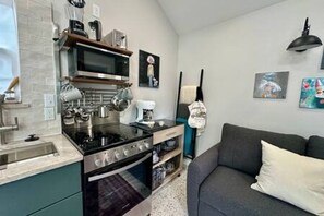 A compact kitchen with a microwave, stove, and oven, a grey sofa, and various kitchen utensils. - New Cottage + Monthly + Fire Pit + Outdoor Kitchen (Dallas)