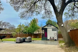 A backyard with a small shed, a covered grill, and a tree. - New Cottage + Monthly + Fire Pit + Outdoor Kitchen (Dallas)