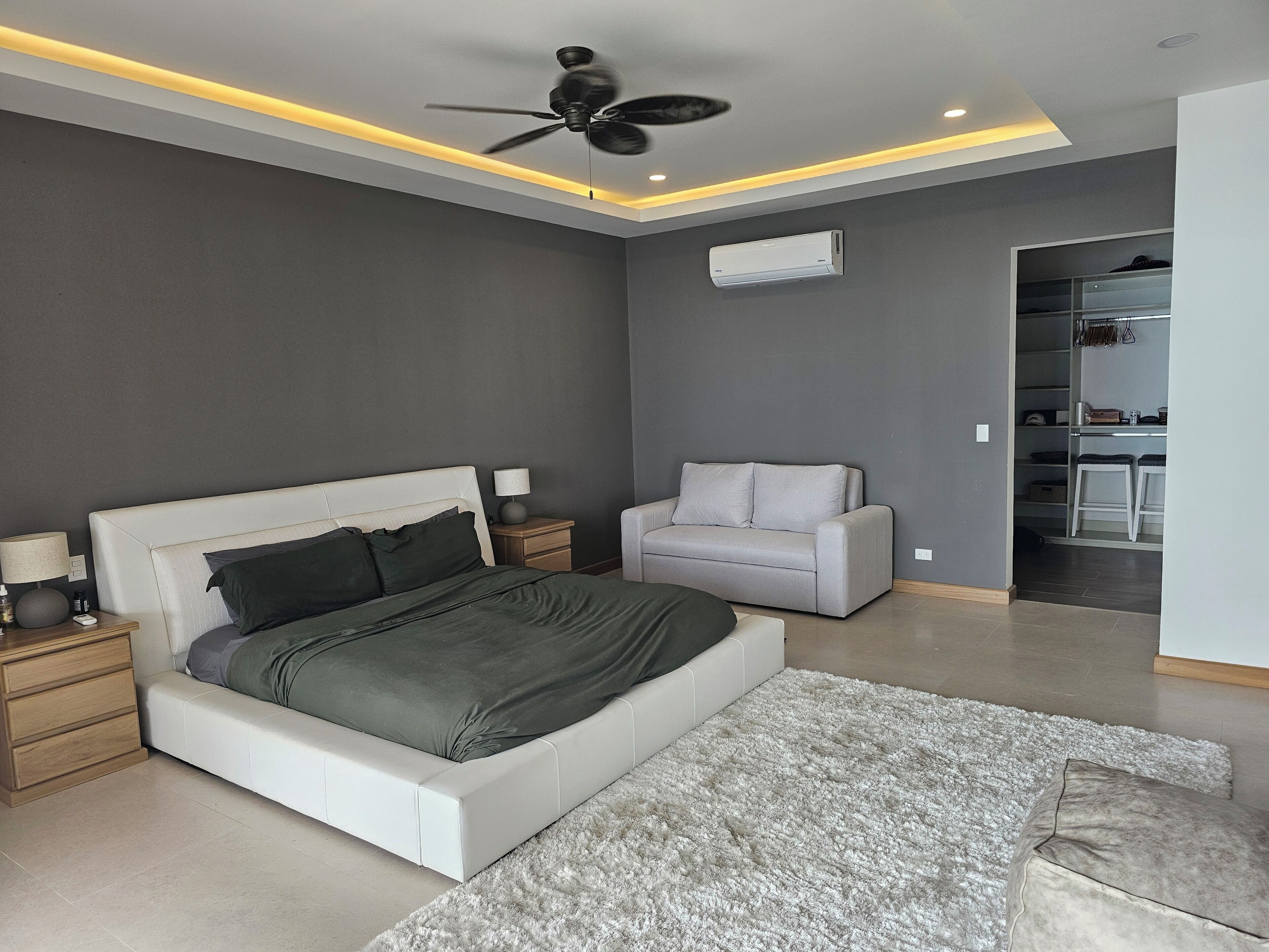 A modern bedroom with a bed, bedside tables, a ceiling fan, an air conditioning unit, and a walk-in closet.