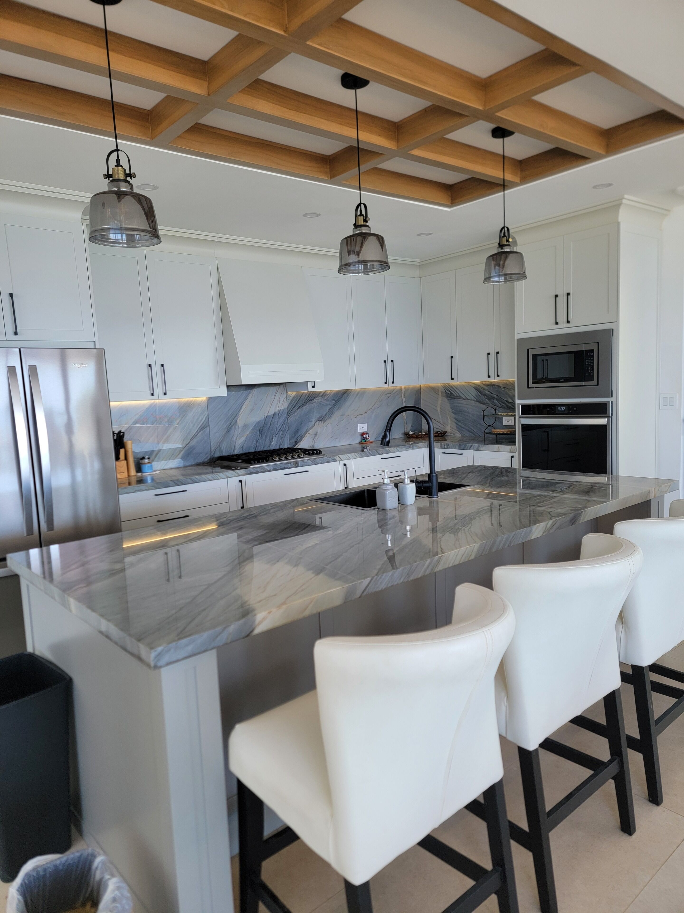 Una cocina moderna con gabinetes blancos, encimera de mármol, electrodomésticos de acero inoxidable e iluminación colgante.