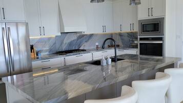 Una cocina moderna con gabinetes blancos, encimera de mármol, electrodomésticos de acero inoxidable e iluminación colgante.