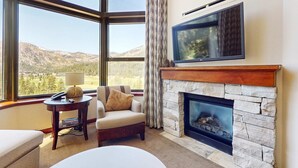 A room with a stone fireplace, a television mounted above it, and a view of a mountainous landscape through large windows. - Everline Resort & Spa Ski-in-ski-out condo with elevator, pool, hot tub, & spa (Olympic Valley)
