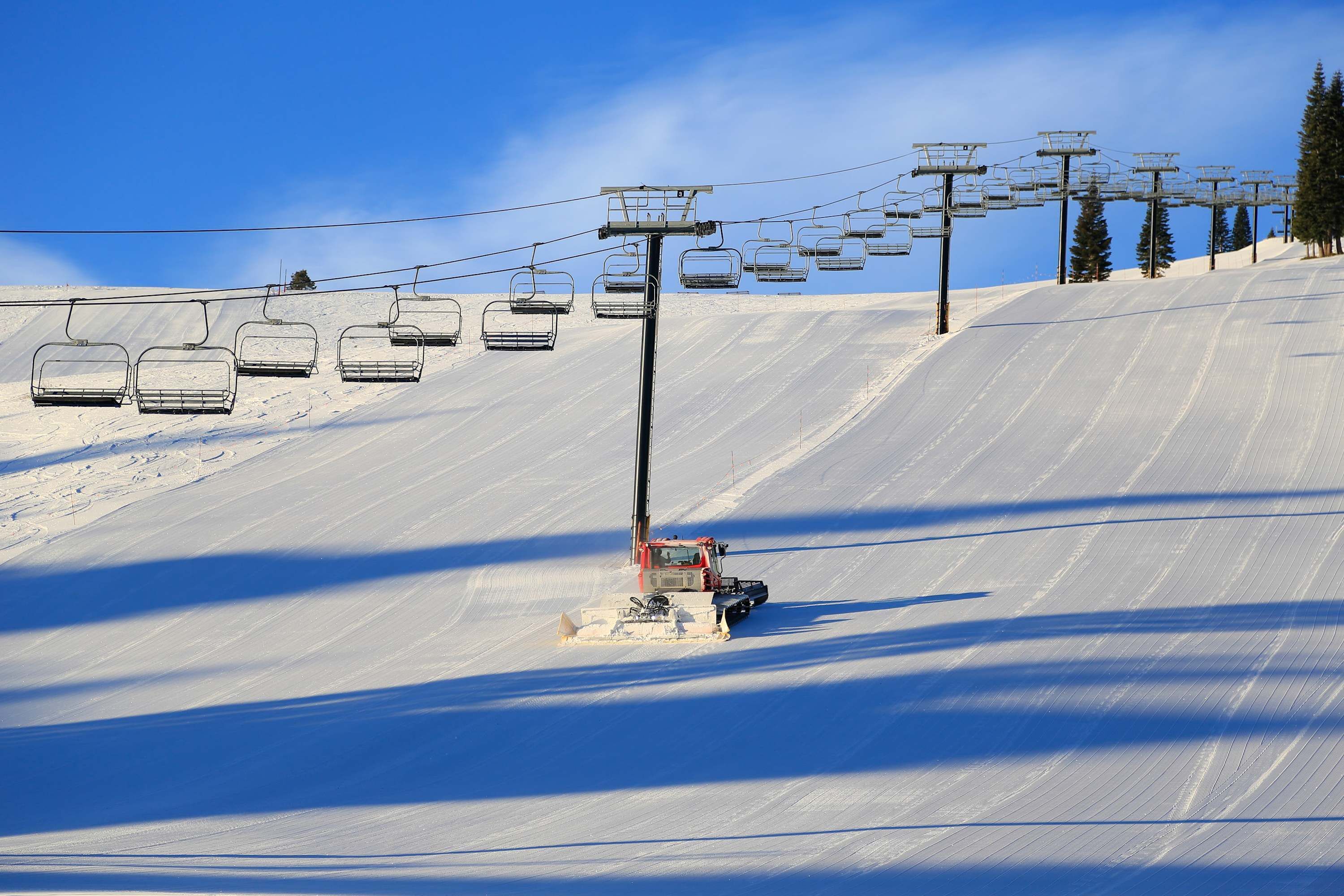 A ski lift with empty chairs, a snow grooming machine, and a clear blue sky.